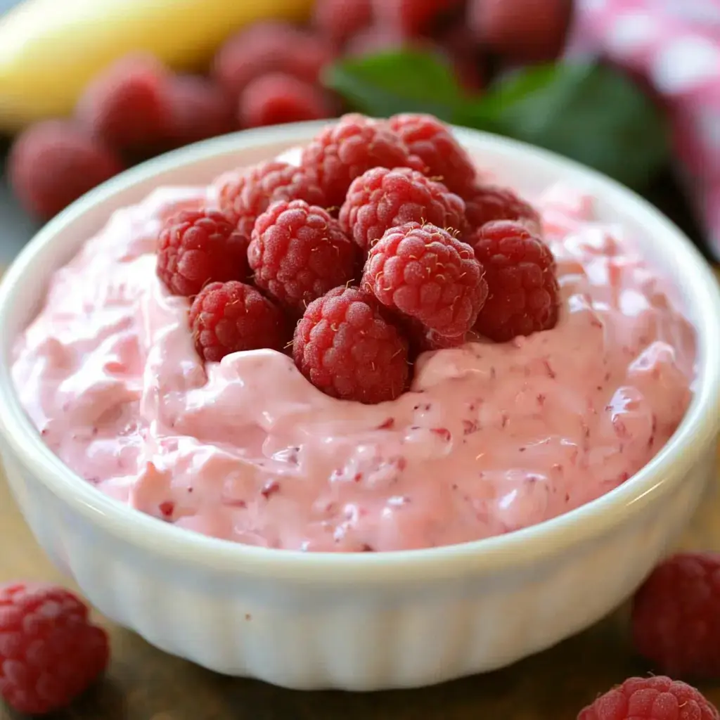 A bowl of pink raspberry yogurt topped with fresh raspberries, surrounded by scattered raspberries and a banana in the background.